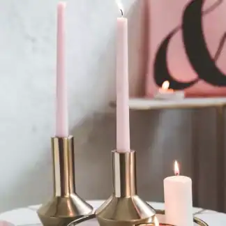 شمعدان ذهبي معدني مودرن بتصميم مخروطي يحمل شمعة طويلة وردية اللون موضوع على طاولة رخامية بجانب شمعة بيضاء.