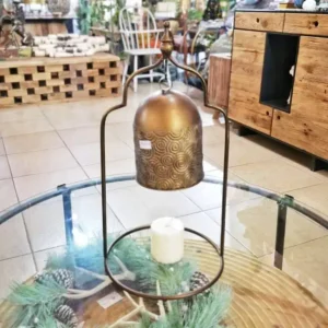 جرس طاولة معدني بلون نحاسي معتق ونقوش حلزونية معلق على إطار معدني موضوع على طاولة زجاجية في متجر ديكور.