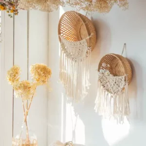 طقم سلات خوص جدارية مكون من قطعتين مزين بمكرمية بيضاء منسدلة معلقة على حائط أبيض بجانب فازة زجاجية وزهور مجففة.