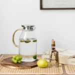 إبريق زجاجي شفاف بمسكة من الراتان الطبيعي وغطاء معدني يحتوي على ماء وليمون موضوع على مائدة بوهيمية بجانب فواكه وأعشاب.