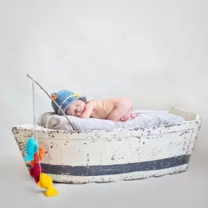 مجسم سفينة خشبية أبيض معتق لتصوير المواليد يظهر بداخله طفل رضيع نائم بجانبه صنارة صيد صغيرة ودمى سمك محاكة