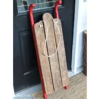 مجسم زحلقة ثلج خشبية ريفي لتصوير المواليد باللون الخشبي الطبيعي مع حواف حمراء وحبل تعليق موضوع بجانب باب أسود