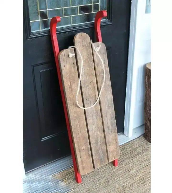 مجسم زحلقة ثلج خشبية ريفي لتصوير المواليد باللون الخشبي الطبيعي مع حواف حمراء وحبل تعليق موضوع بجانب باب أسود