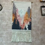 مكرمية تعليقة جدارية يدوية بمشهد مطبوع لغابة خريفية بألوان دافئة مع شراريب مكرمية بيضاء معلقة في ركن منزلي هادئ