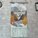 مكرمية جدارية لوحة قماشية بمشهد مطبوع لطريق يمر بين الجبال وشمس الغروب مع شراريب مكرمية بيضاء يدوية معلقة في ركن فني