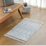 دواسة قماشية مودرن باللون الرمادي المزرق مع أهداب جانبية موضوعة في زاوية استرخاء عصرية بجانب مقعد خشبي وكتب