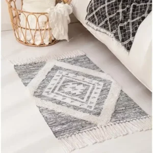 دعاسات باب البيت قماشية بنقوش هندسية باللون الرمادي والأبيض مع أهداب جانبية موضوعة في مدخل عصري بجانب سلة قش ونباتات