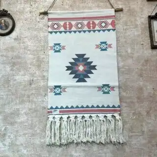 مكرمية بيضاء جدارية يدوية بنقوش هندسية بألوان الأزرق والوردي مع شراريب قطنية كثيفة معلقة في مدخل منزل عصري