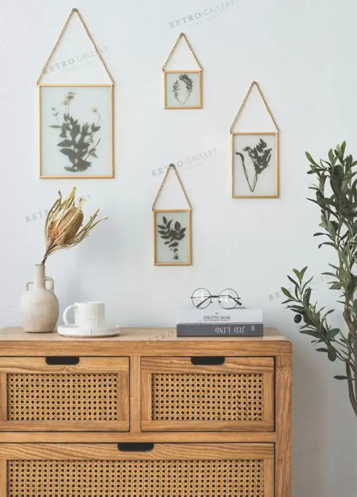 مجموعة لوحات زجاجية بإطارات ذهبية وسلاسل تعليق تضم أوراقاً نباتية مضغوطة معلقة على حائط أبيض فوق خزانة خشبية بتنسيق بوهيمي.